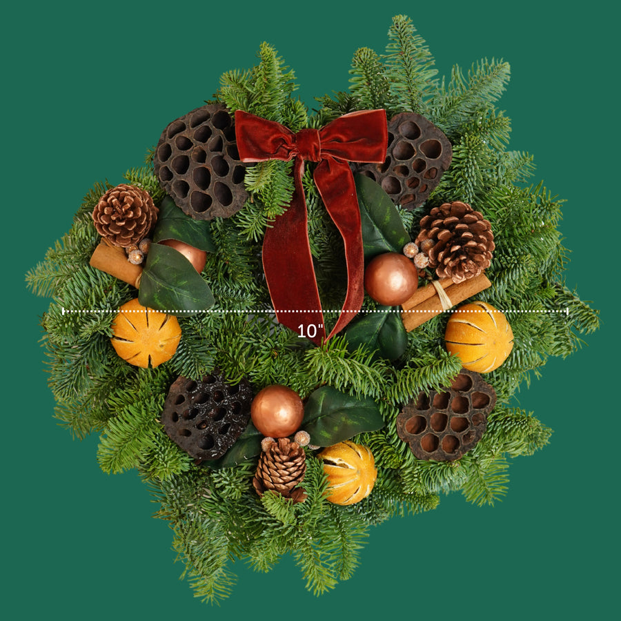 Cinnamon Spice Christmas Wreath A cinnamon spice wreath displayed against a green background, featuring a red velvet bow, pinecones, dried oranges, cinnamon sticks, lotus pods, and copper baubles, with a measurement line showing a 10-inch width. 3