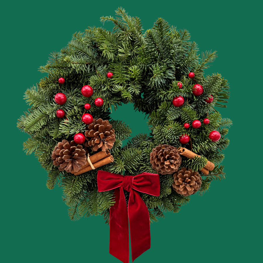 Festive Red Christmas Wreath Front view of the Festive Red wreath featuring evergreen foliage, glossy red berries, pinecones, and cinnamon sticks tied with a velvet bow, set against a plain green background. 3