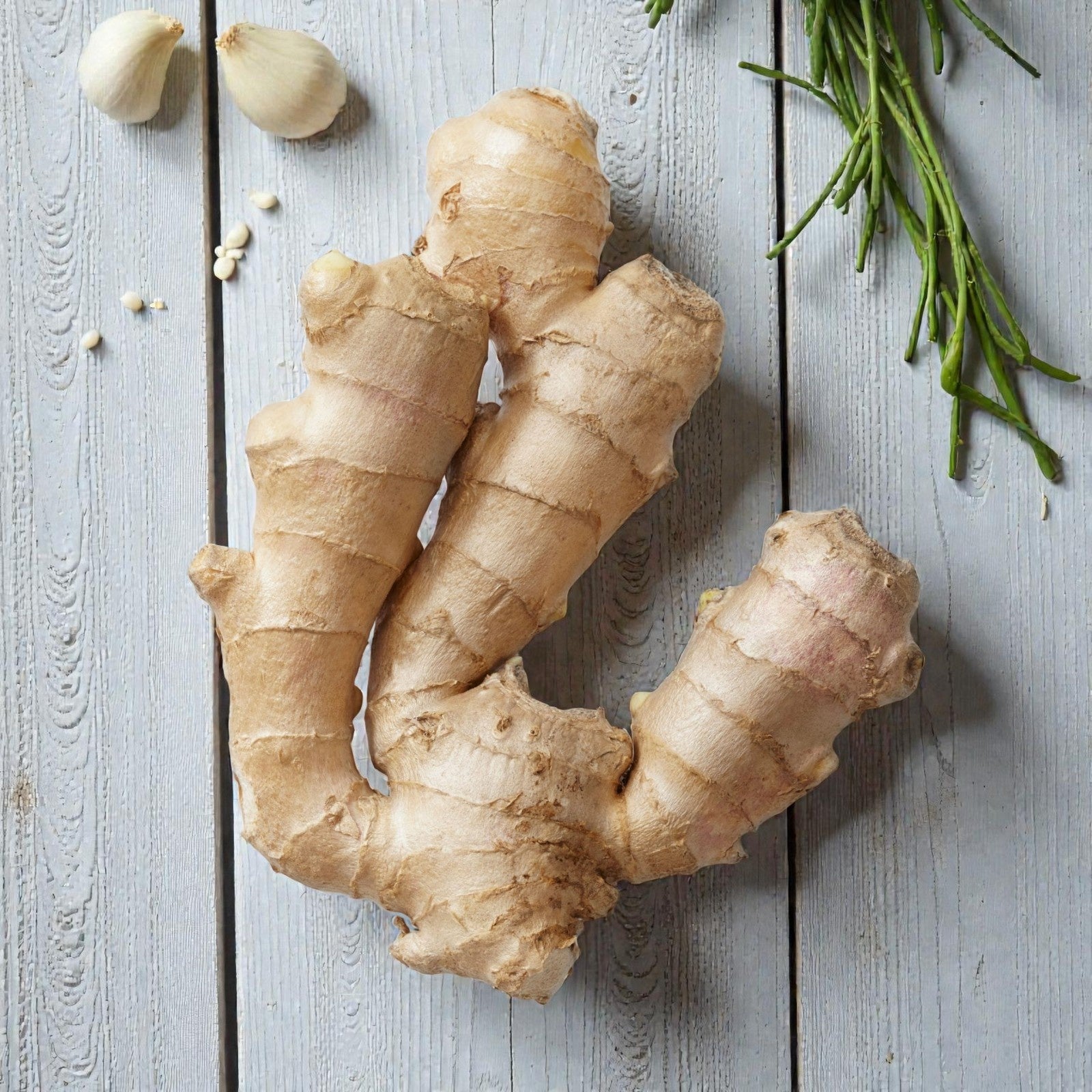 A fresh ginger rhizome with smooth tan skin displayed on a light wooden surface, surrounded by garlic cloves and herbs.