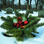 Christmas Grave Posy A small festive grave posy arrangement made from evergreen fir foliage, decorated with red berries, pinecones, green leaves and a red bauble, placed outdoors on a snowy surface in a cemetery. 1