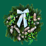 Mistletoe Kisses Christmas Wreath Front view of the Mistletoe Kisses Christmas wreath on a green background, showing a 10-inch diameter with decorative pinecones, white berries, and silver bow detail. 3