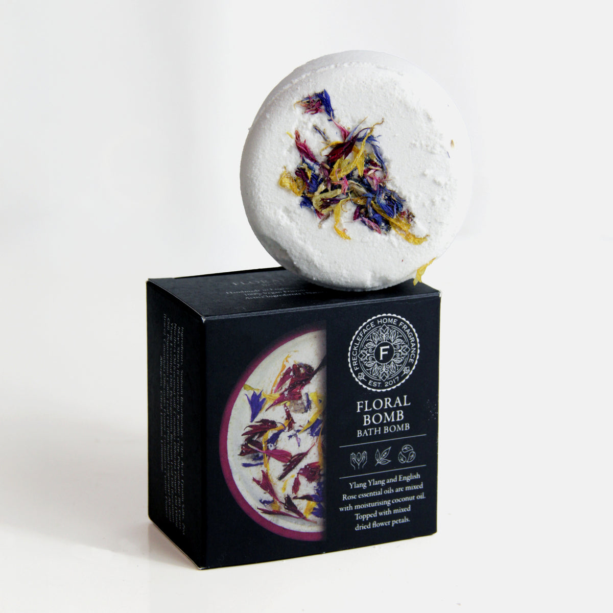 Freckleface Floral Bomb Bath Bomb White round bath bomb topped with colourful dried flower petals, displayed on its black and purple box labelled “Floral Bomb Bath Bomb.” The packaging mentions ylang ylang and English rose essential oils. 1