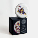 Freckleface Floral Bomb Bath Bomb White round bath bomb topped with colourful dried flower petals, displayed on its black and purple box labelled “Floral Bomb Bath Bomb.” The packaging mentions ylang ylang and English rose essential oils. 1