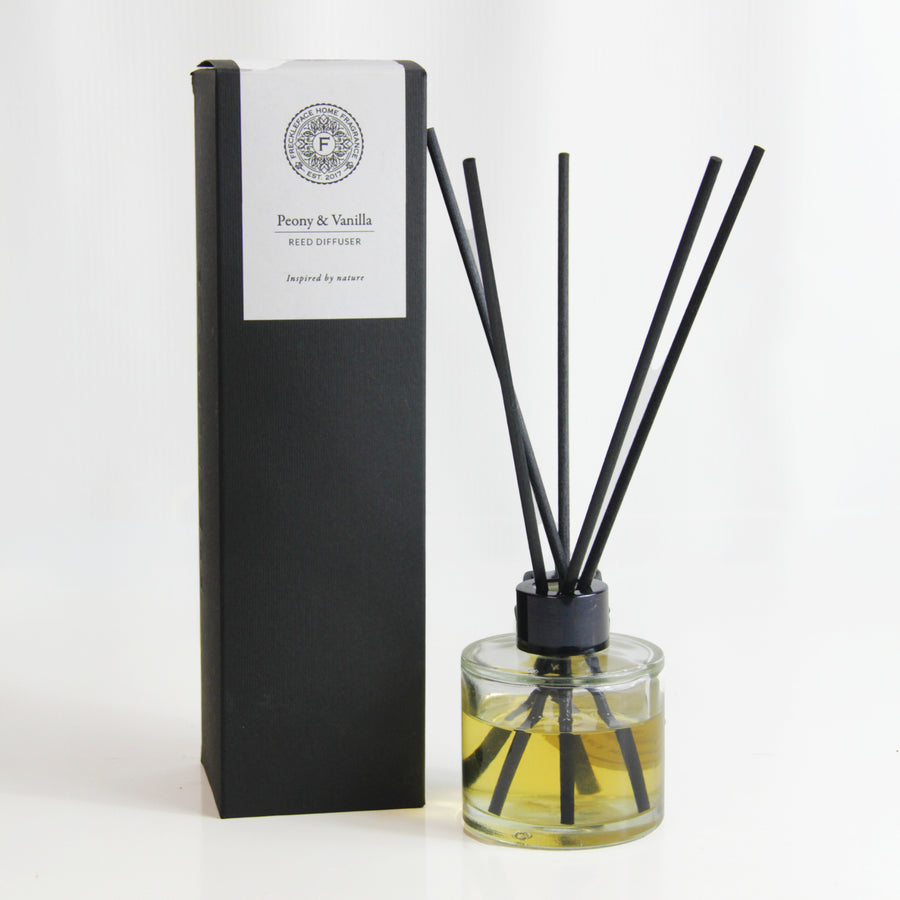 Freckleface Peony & Vanilla 125ml Reed Diffuser Peony and Vanilla reed diffuser with black reeds in a clear glass bottle filled with golden liquid, placed beside its tall black box with white label. 1