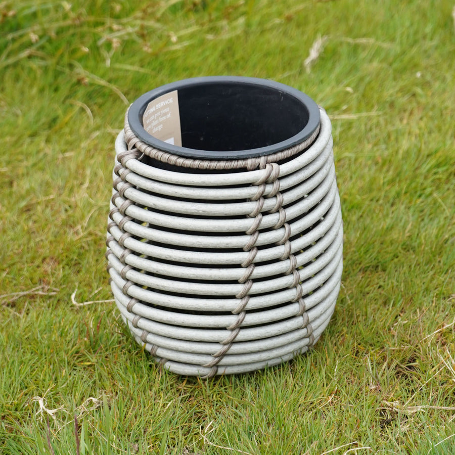 Mica Plant Pots - White Cane Small white round rattan-style planter with black plastic inner pot, placed on grass. 1